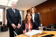 Die Botschafterin sitzt am Besprechungstisch im Präsidentinnenzimmer und trägt sich in das vor ihr liegende Gästebuch ein. Der Honorarkonsul und die Bürgerschaftspräsidentin stehen neben ihr. Alle gucken freundlich in die Kamera.