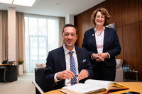 Der Botschafter sitzt am Konferenztisch im Präsidentinnenzimmer und trägt sich in das vor ihm liegende Gästebuch ein. Er trägt einen dunklen Anzug mit gestreifter Krawatte. Antje Grotheer steht neben ihm. Beide gucken freundlich in die Kamera.