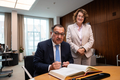Der Botschafter sitzt am Konferenztisch im Präsidentinnenzimmer und trägt sich in das vor ihn liegende Gästebuch ein. Die Präsidentin steht neben ihm. Beide gucken freundlich in die Kamera.