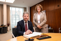 Der Botschafter sitzt am Besprechungstisch im Präsidentinnenzimmer und trägt sich in das vor ihm liegende Gästebuch ein. Die Präsidentin steht neben ihm. Beide gucken freundlich in die Kamera.
