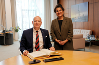 Der Generalkonsul sitzt am Konferenztisch im Präsidentinnenzimmer und trägt sich in das vor ihm liegende Gästebuch ein. Die Vizepräsidentin steht neben ihm. Beide gucken freundlich in die Kamera.