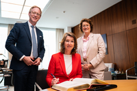 Die Botschafterin sitzt am Konferenztisch im Präsidentinnenzimmer und trägt sich in das vor ihr liegende Gästebuch ein. Die Präsidentin und der Honorarkonsul stehen neben ihr. Alle gucken freundlich in die Kamera.