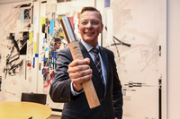 Präsident Imhoff steht in seinem Büro. Er hält den Staffelstab in die Kamera und lächelt.