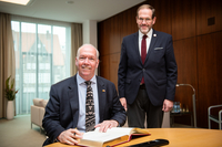 Der Botschafter sitzt am Konferenztisch im Präsidentenzimmer und trägt sich in das Gästebucht ein. Claas Rohmeyer steht neben ihm, beide tragen einen Anzug und gucken freundlich in die Kamera.