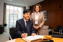 Lin Dong sitzt am Besprechungstisch im Präsidentinnenzimmer und trägt sich in der vor ihm liegende Gästebuch ein. Antje Grotheer steht neben ihm. Beide schauen freundlich in die Kamera.