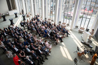 Ein Blick von der Empore in den Festsaal. Die Menschen sitzen in Stuhlreihen und hören der Rednerin zu. Neben dem Rednerpult liegen die Instrumente der Musiker.